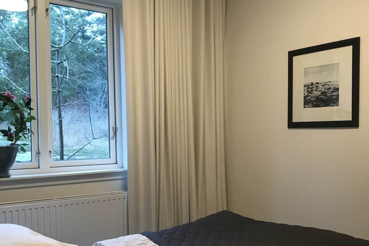 Ein Schlafzimmer mit Bett, einem Fenster mit Vorhängen, einem Heizkörper, einem gerahmten Bild an der Wand und einem Blumentopf am Fenster.