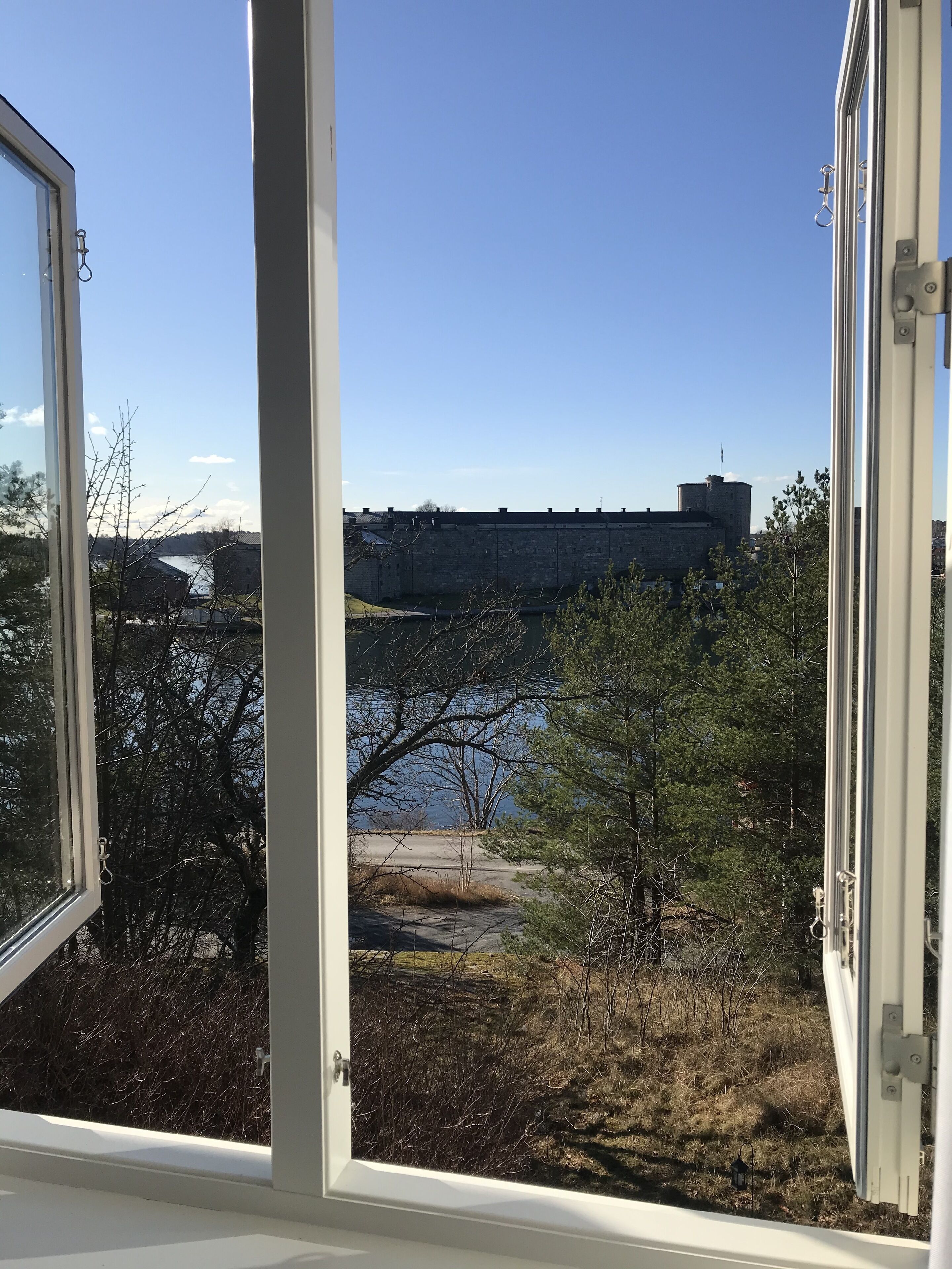 Vista a través de una ventana de un edificio de piedra, con cielo despejado, una masa de agua y algunos árboles.