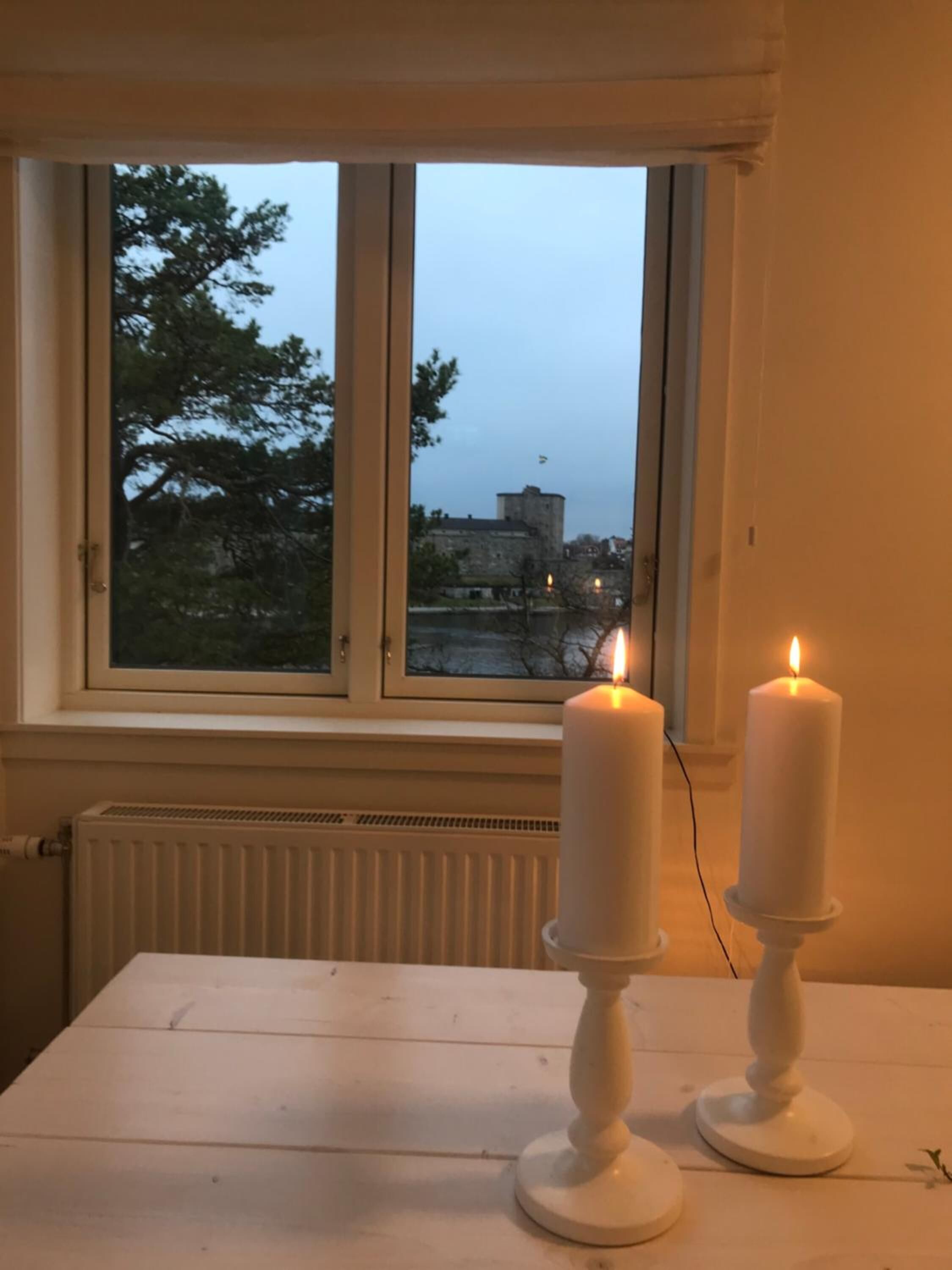 Una habitación con una mesa blanca y dos velas blancas en candelabros. Se ve una ventana con vistas a árboles y un edificio.