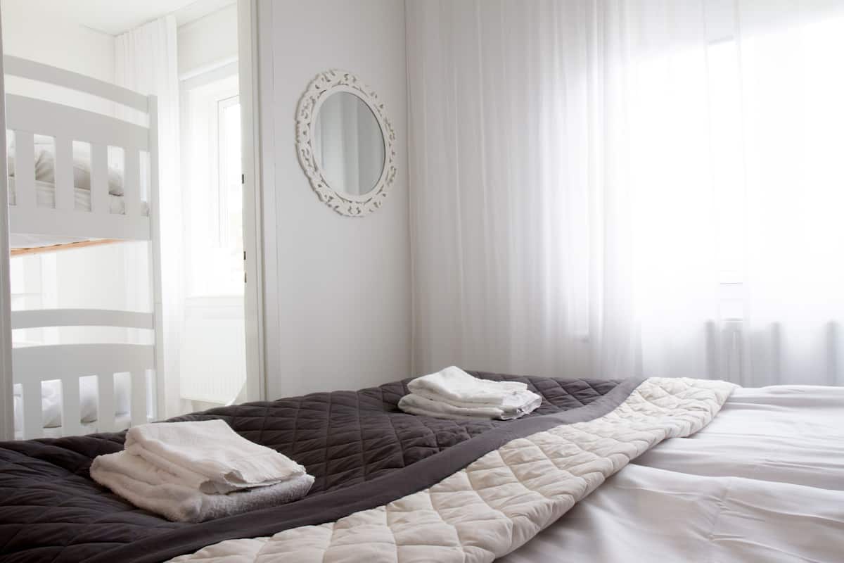 Ein ordentlich bezogenes Bett mit einer gesteppten Decke und gefalteten Handtüchern, ein weißes Babybett und ein wandmontierter Spiegel.