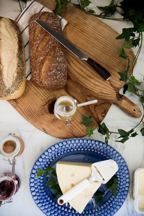 Tábua de madeira com pão, faca, um frasco de mel, um prato azul com queijo e um cortador de queijo, e uma garrafa de vinho.