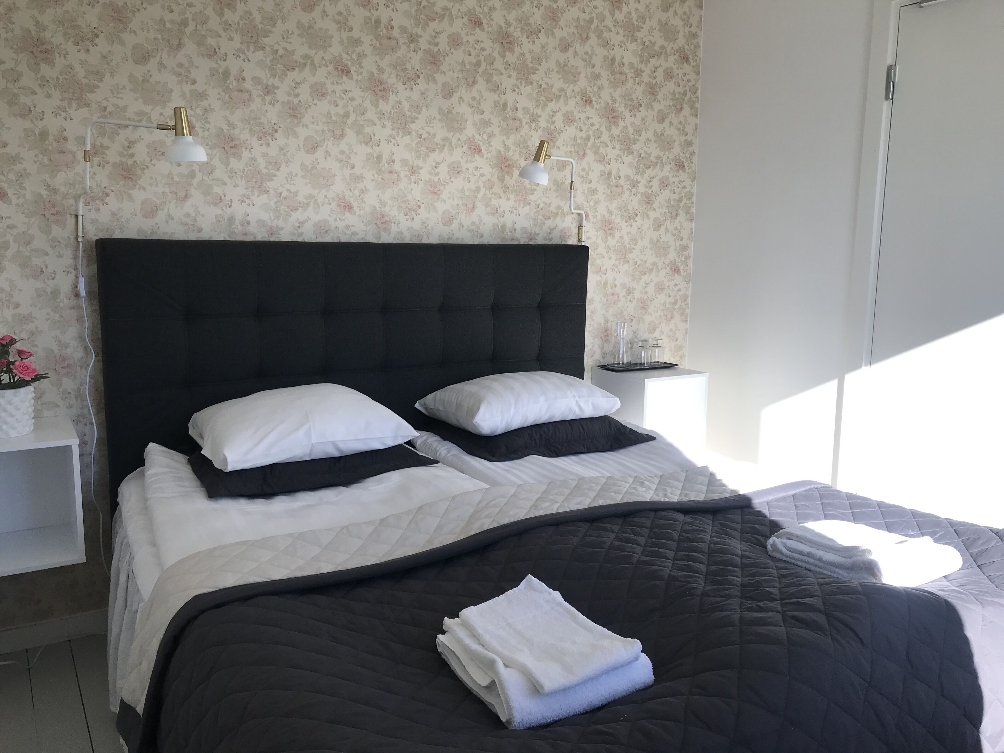A neatly made bed with a black headboard, white and black pillows, and a folded white towel on the bed. There are two bedside tables with lamps, a white wall with floral wallpaper, and a white door in the background.