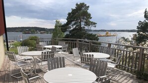 A deck with round tables and chairs, overlooking a body of water with boats and a distant shoreline.