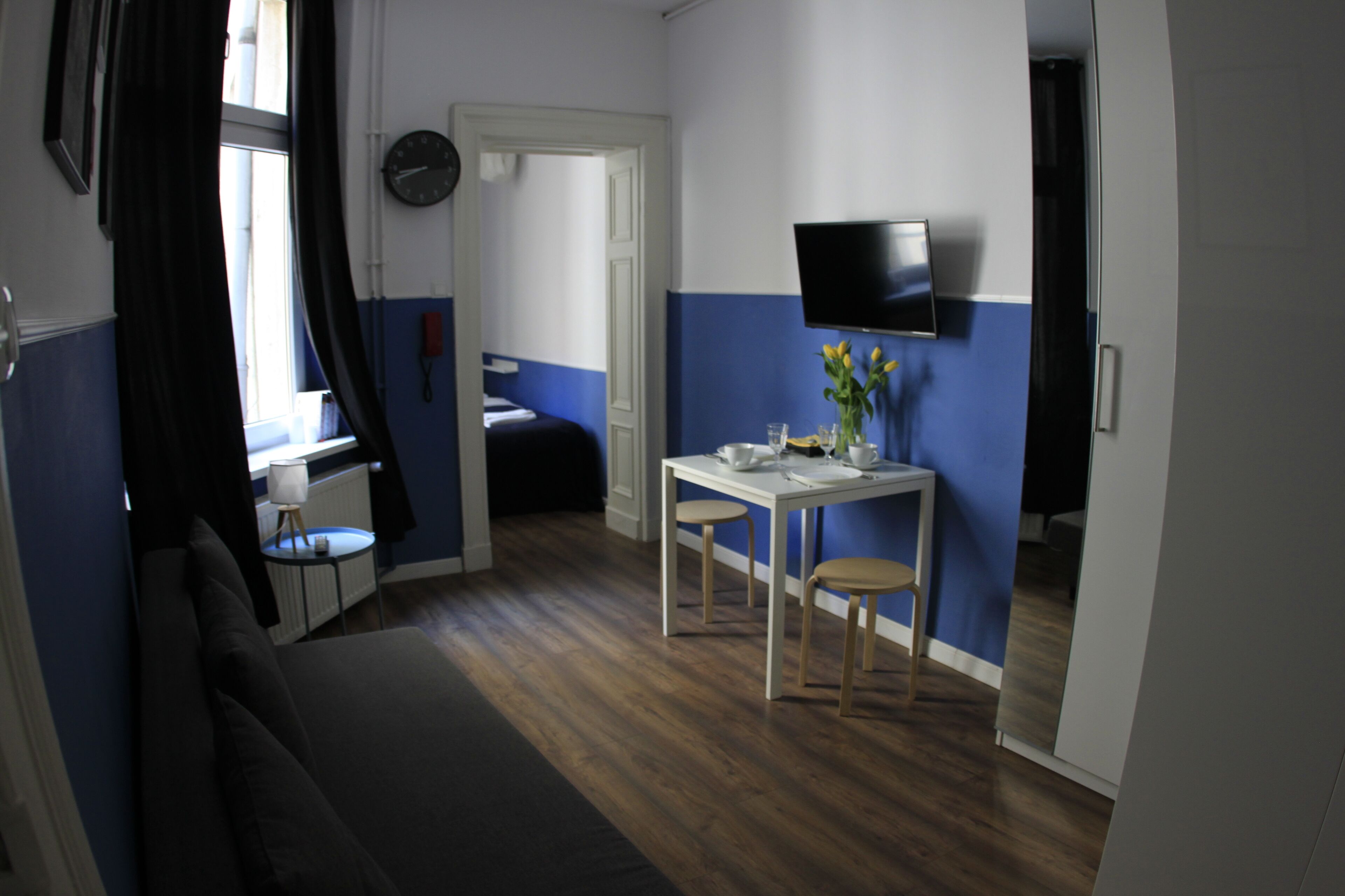 Habitación compacta con cama, una pequeña mesa con sillas, un televisor en la pared y una ventana con cortinas.