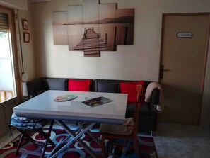 A room with a white table, a black sofa with red cushions, a wooden door labeled 'CHAMBRE', and a wall with a framed artwork depicting a dock and mountains.