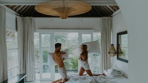 Une chambre avec un lit, un banc, un ventilateur de plafond et une vue sur la verdure à l’extérieur.