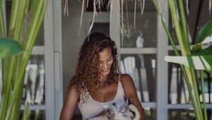Une personne tenant un chat dans une pièce dont le sol est en bois et où se trouvent de grandes plantes vertes.
