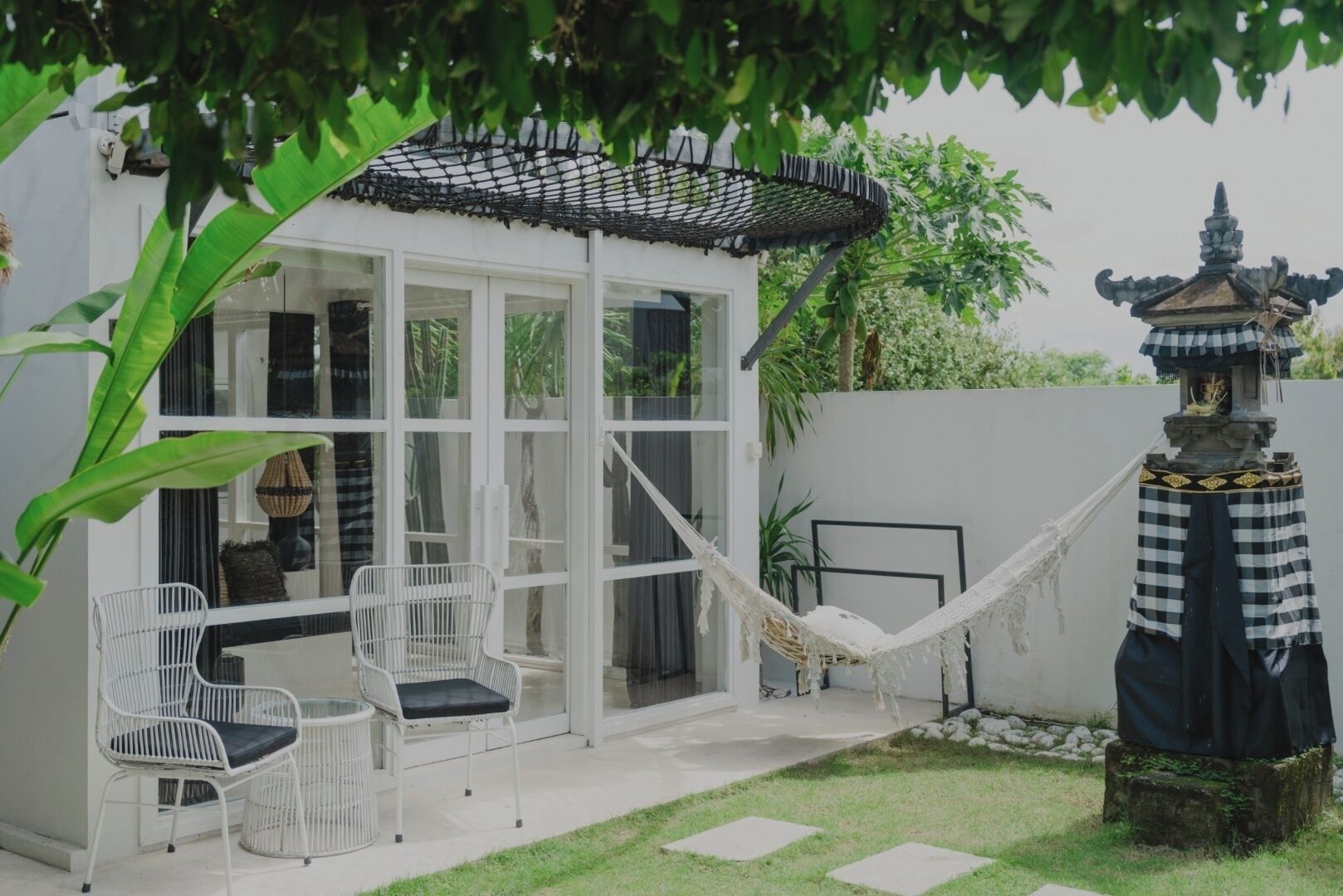 Una casa moderna con puerta de vidrio, mobiliario de mimbre blanco, una hamaca y una estatua tradicional.