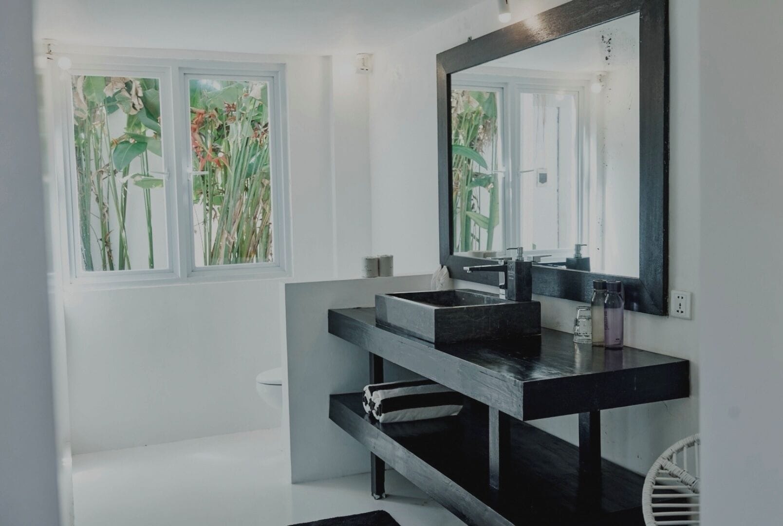 Ein modernes Badezimmer mit einem schwarzen Waschbecken, einem großen Spiegel und einem Fenster mit Blick auf Pflanzen.