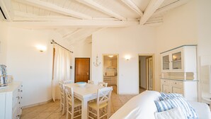 Un interior luminoso y blanco con comedor, cocina y una cama con almohadones a rayas.