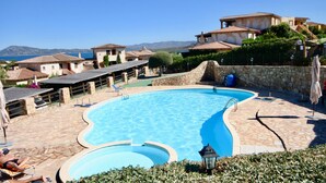 Una piscina con plataforma de piedra, rodeada de edificios de arquitectura mediterránea, y una vista despejada al mar en el fondo.