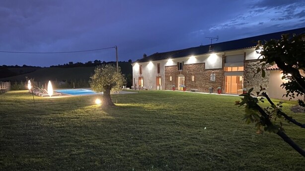 Une propriété résidentielle bien éclairée, dotée d’une piscine, d’une pelouse et d’un bâtiment en pierre avec de nombreuses fenêtres et portes.