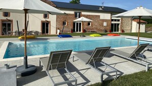 Une piscine entourée de chaises longues et de parasols, elle-même bordée d’une maison en briques surmontée d’un toit en tuiles.