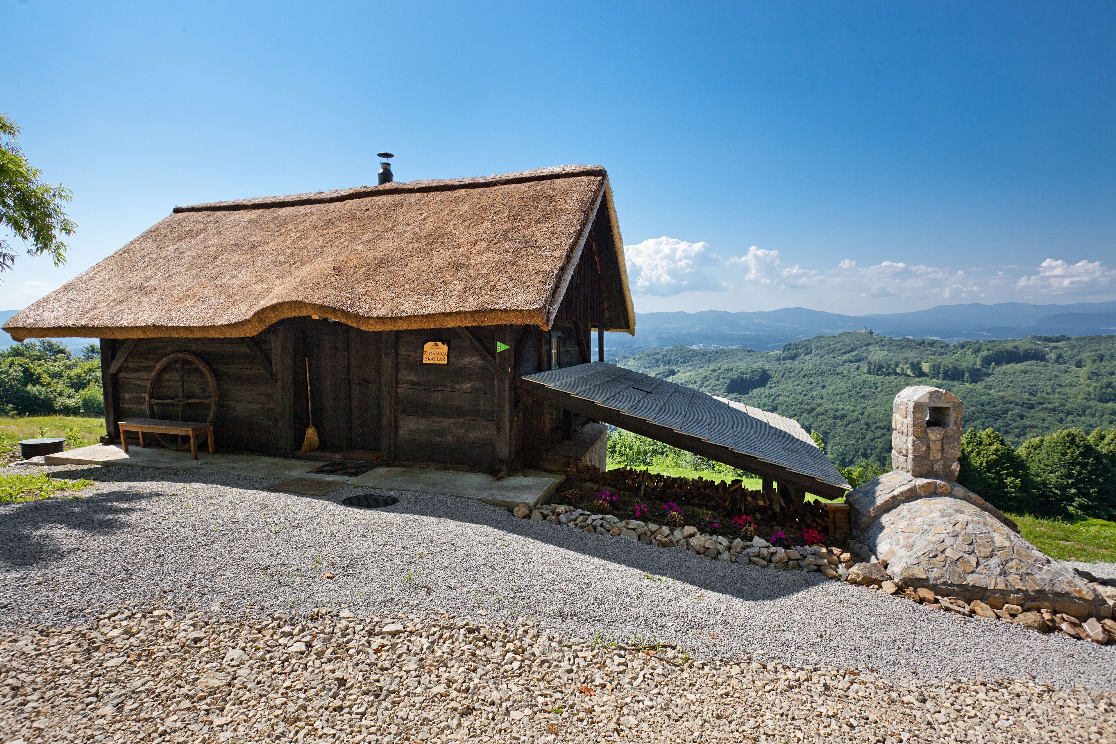 A rustic wooden cabin with a thatched roof, a stone chimney, and a gravel driveway.
