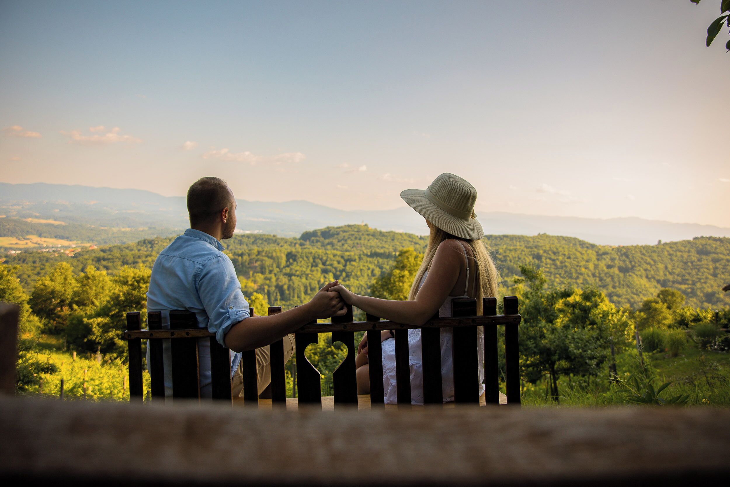 A couple sitting on a bench overlooking a scenic landscape with hills and trees.
