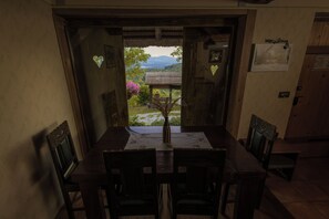 Un comedor con mesa y sillas, vistas a la vegetación y montañas a través de un ventanal abierto, e interior rústico con paredes de madera y estructura de techo de paja.