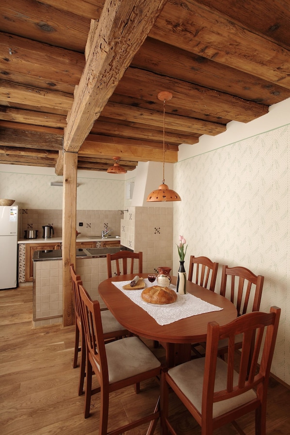 Un coin repas rustique avec une table et des chaises en bois, une cuisine en arrière-plan et un plafond avec des poutres apparentes.