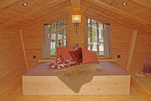 Une chambre dans une cabane en bois, avec un lit, deux fenêtres et un luminaire au plafond.