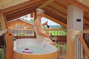 A woman in a white bathrobe sitting in a wooden hot tub with flowers and candles, overlooking a wooden deck and a view of a house and trees. - Chalet Ochkazl für 1-3 Personen (46qm) (Bernau am Chiemsee)