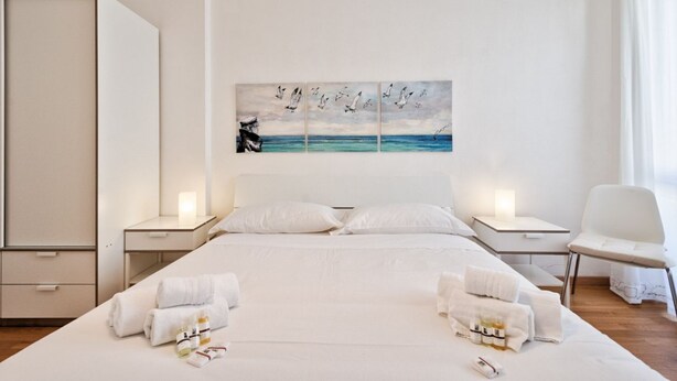 A hotel room with a large bed, two bedside tables, a chair, and a painting of birds flying over the sea.