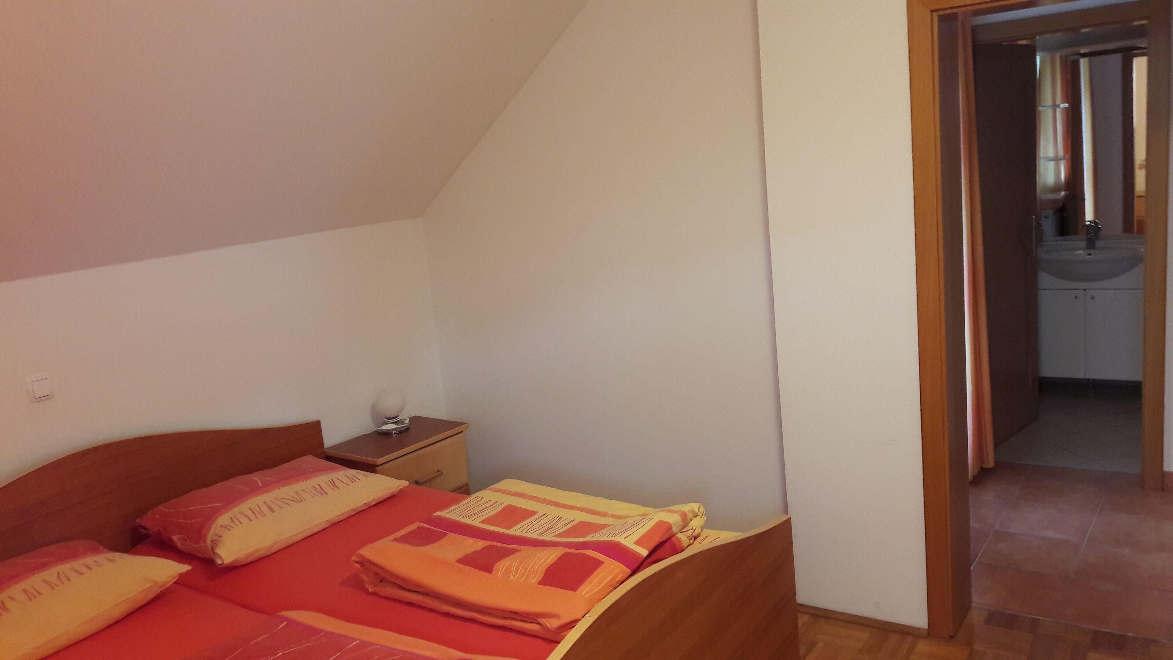 Un dormitorio con una cama de madera, una mesita de noche y un baño visible a través de una puerta abierta.