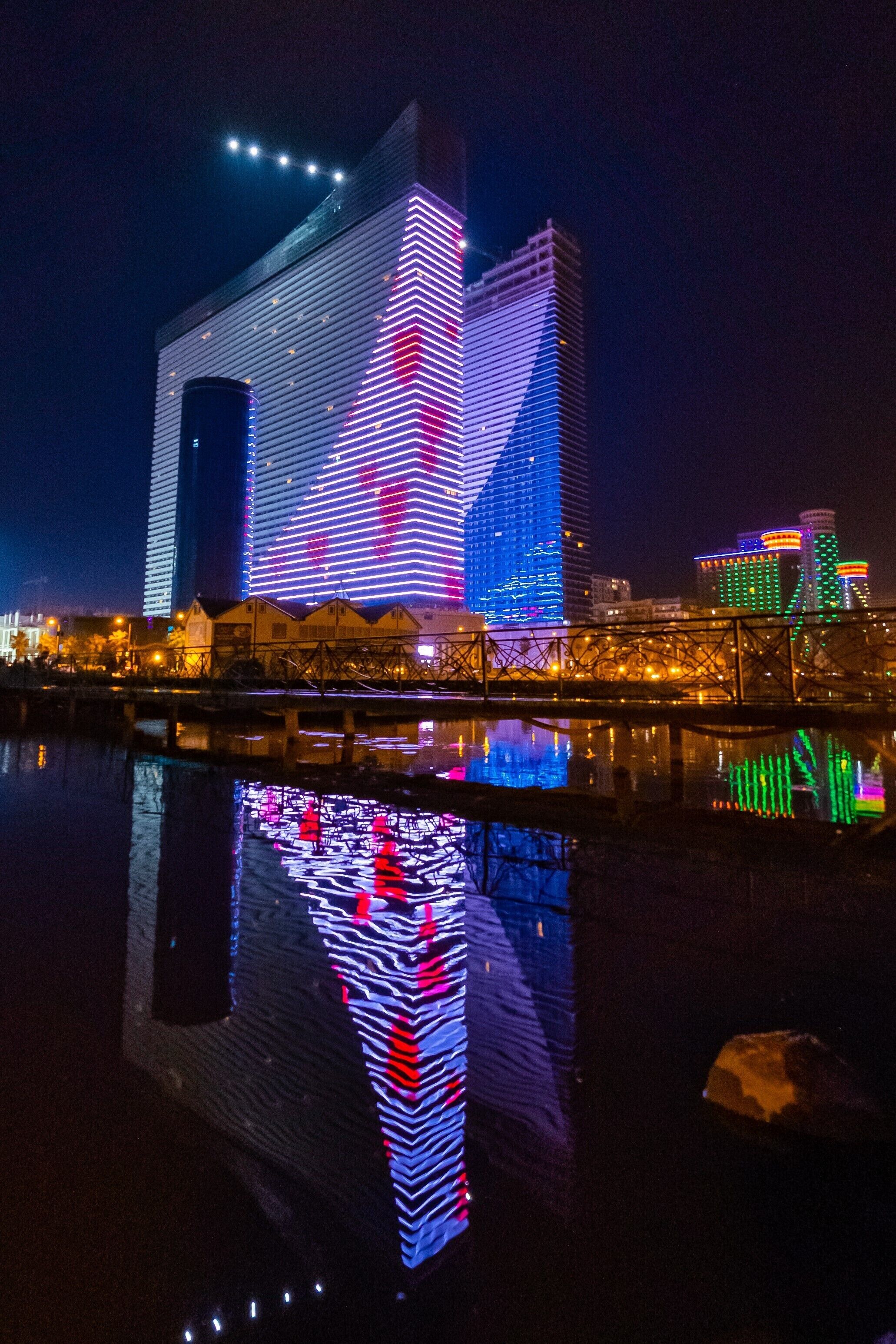 Une vue nocturne d’une ville, avec des bâtiments illuminés se reflétant dans une étendue d’eau.
