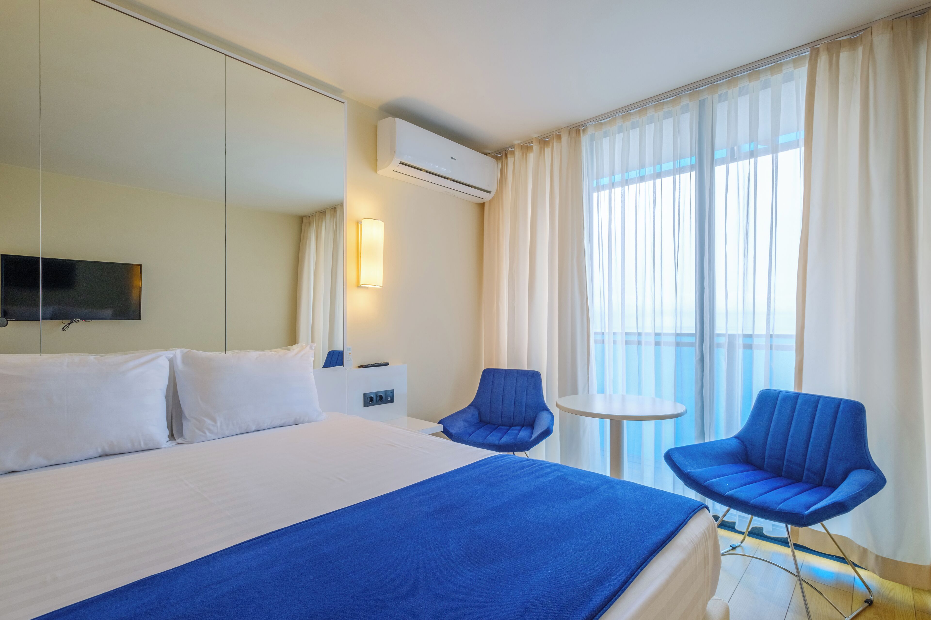 A hotel room with a large bed, two blue chairs, a small table, a TV, and an air conditioning unit.