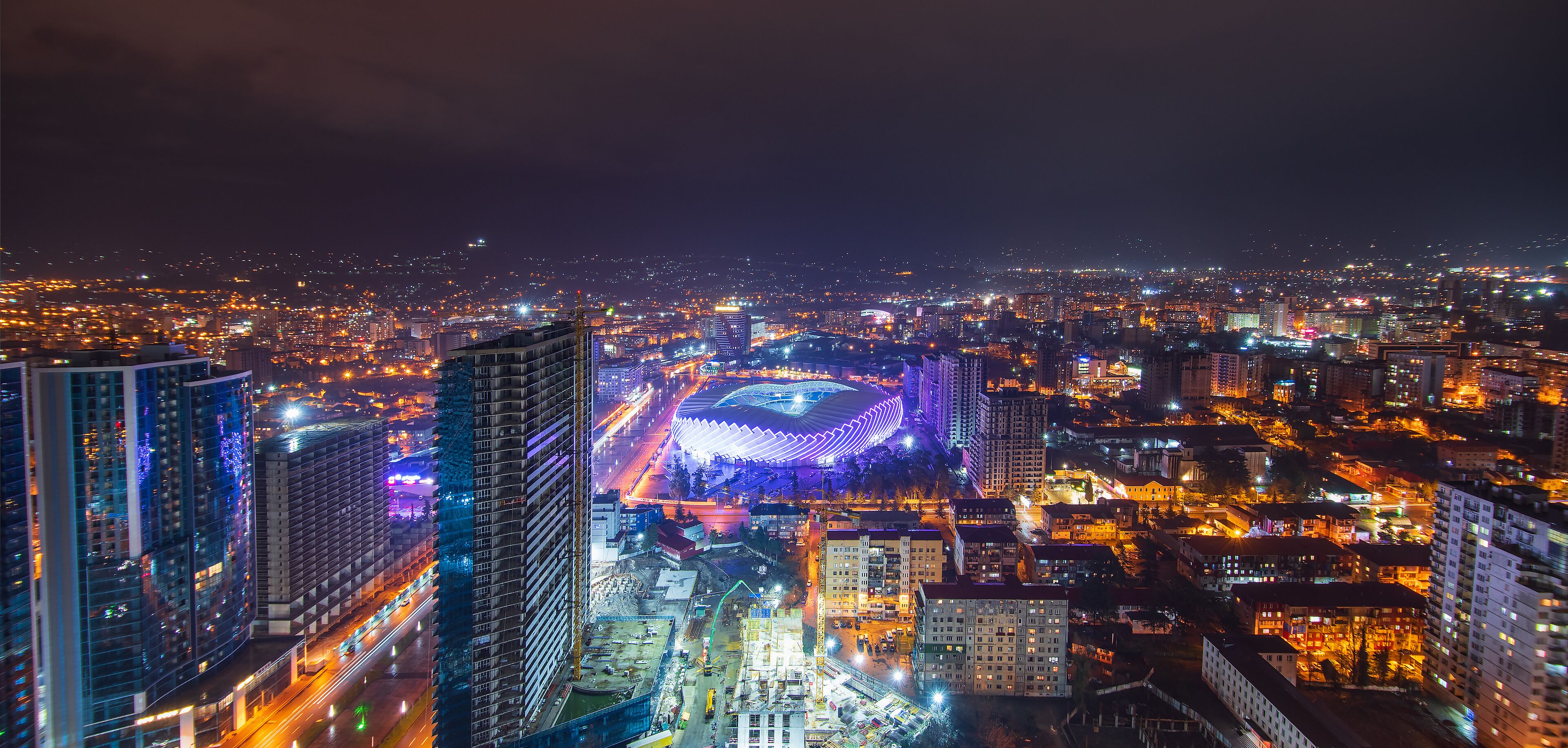 A nighttime cityscape with illuminated buildings and a stadium with a distinctive roof structure.