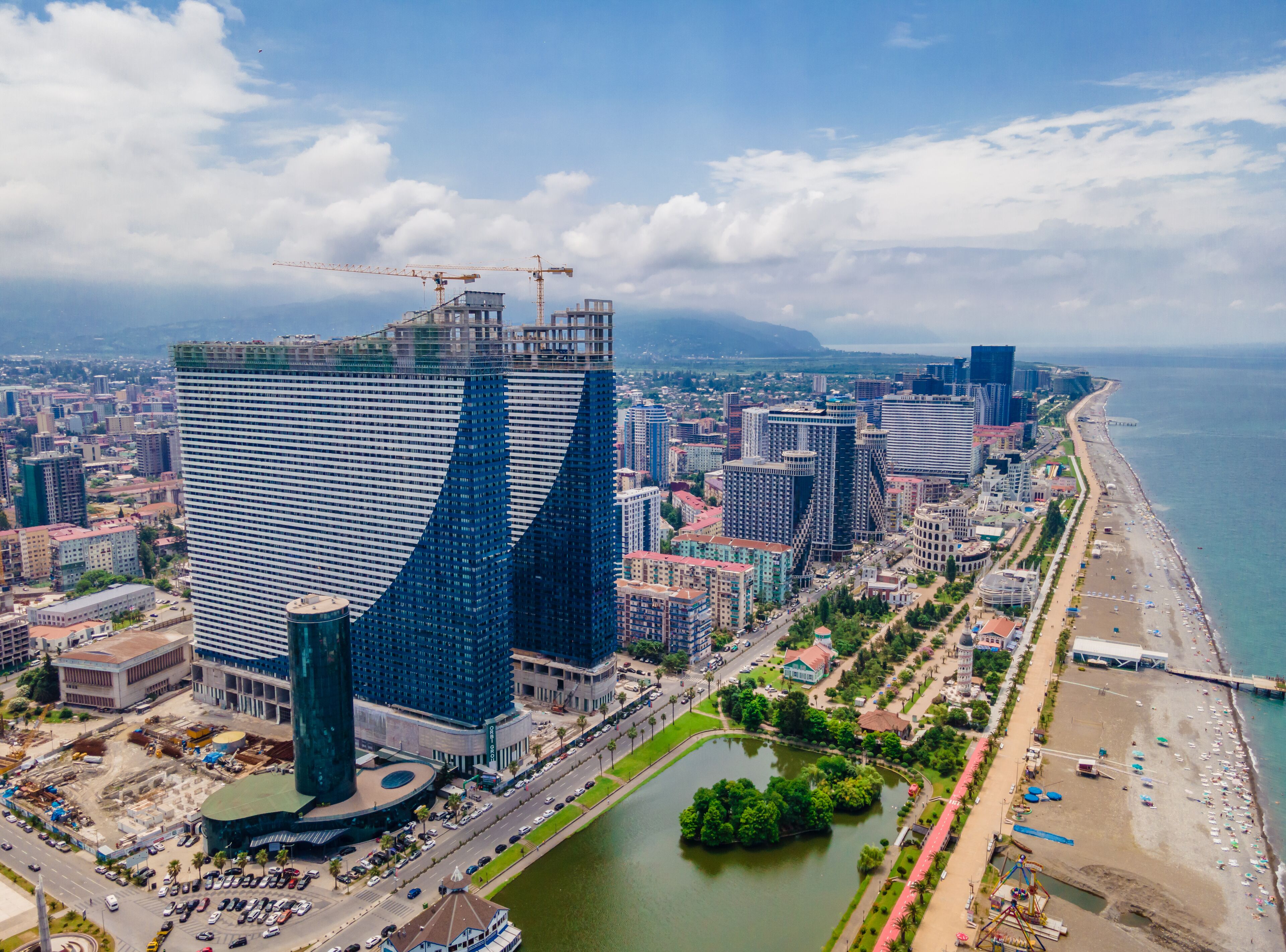 A modern high-rise hotel with a unique curved design, situated near a waterfront with a beach and a river.