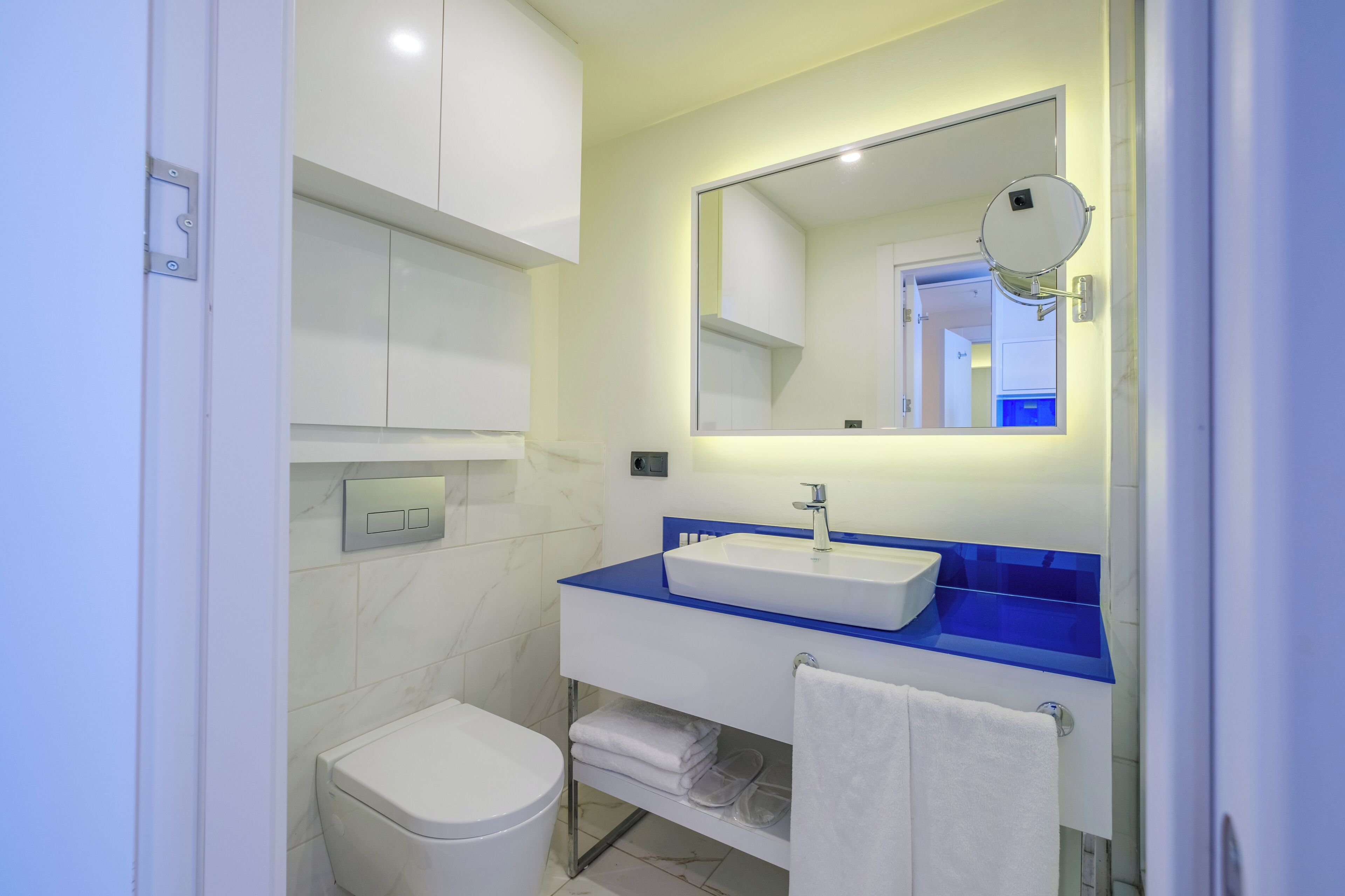 A modern bathroom with a white toilet, a sink with a blue countertop, and a mirror with a round magnifying glass.