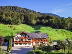 A two-story lodge with a sloped roof, surrounded by lush green hills and a forested mountain backdrop.