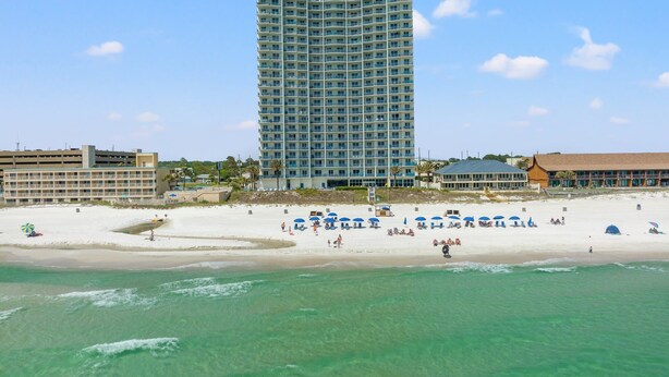 A beachfront hotel with multiple floors, a sandy beach with umbrellas, and a clear view of the ocean.