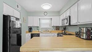 Une cuisine avec des meubles blancs, un réfrigérateur noir, un plan de travail en bois, un évier et une horloge au mur.