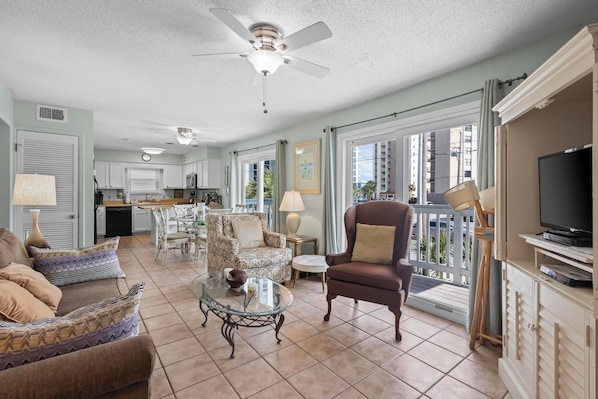 A spacious living area with a ceiling fan, a dining table with chairs, a sofa, a TV, and a balcony with a railing.