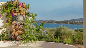 A stone wall with a metal bell, surrounded by flowering plants and overlooking a coastal landscape. - Casa di Nonna Anna (Olbia)