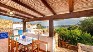 A covered outdoor dining area with a stone wall, a table with chairs, and a view of the sea. - Casa di Nonna Anna (Olbia)