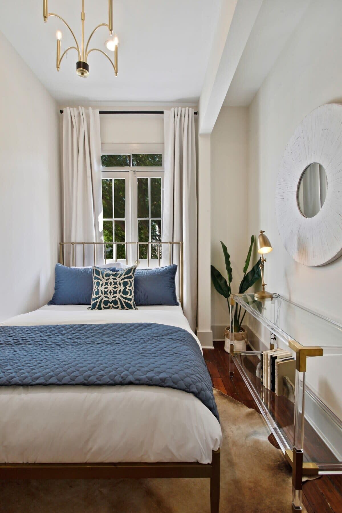A bedroom with a large bed, a bedside table with a lamp, a round mirror on the wall, and a window with curtains.
