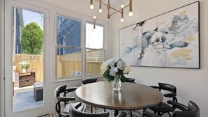 A dining area with a round table, black chairs, a vase with flowers, a large abstract painting, and a view of a wooden fence and trees through large windows.