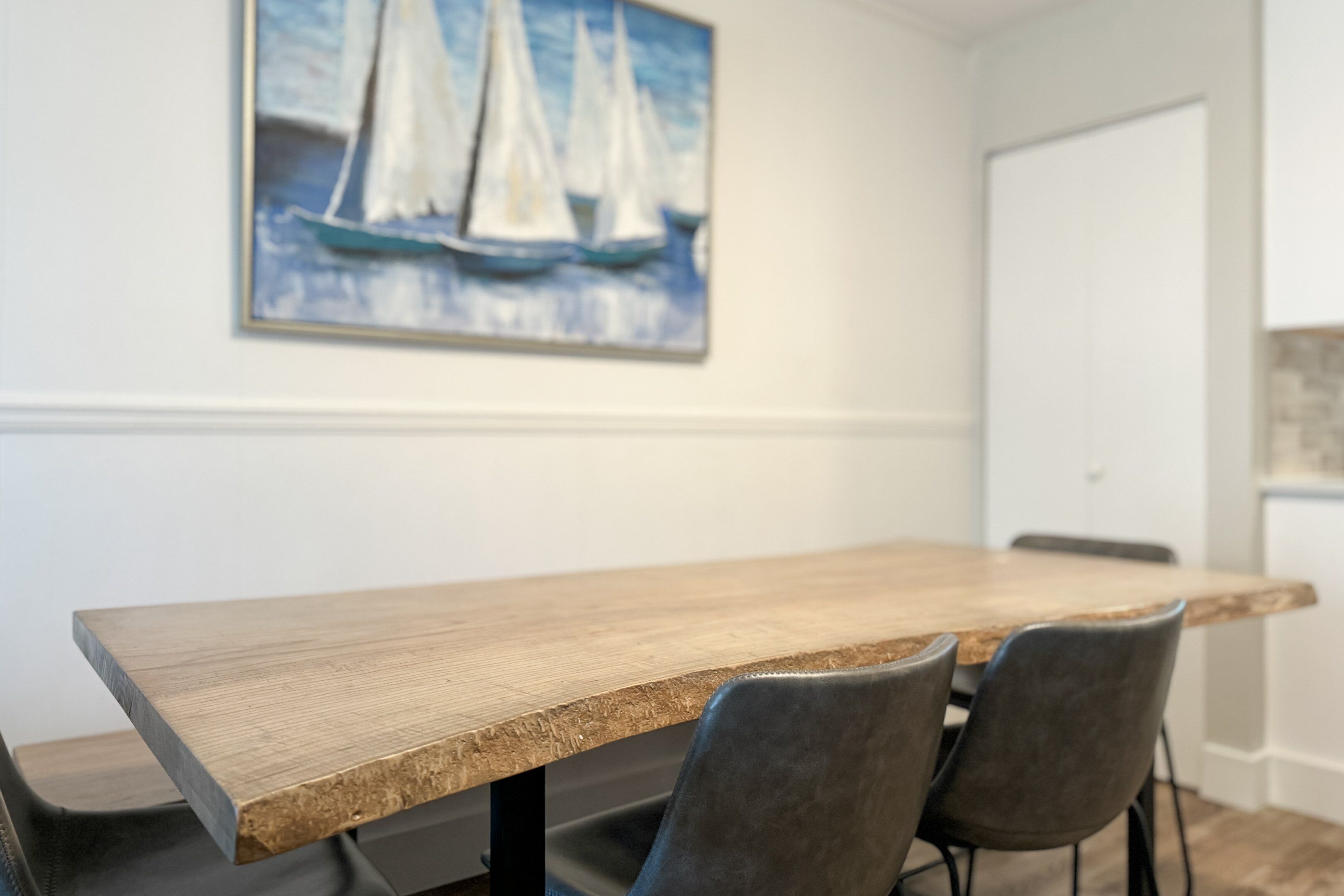 Un comedor con una mesa y sillas de madera, un cuadro de veleros en la pared y una puerta blanca al fondo.