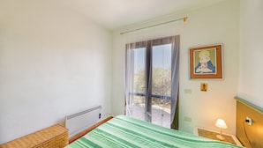 A hotel room with a bed, a radiator, a window with curtains, a framed picture on the wall, and a bedside lamp. - Casa Girgolu (Loiri Porto San Paolo)