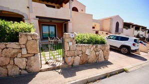 A stone wall with a gate, a white car parked in front, and a building with a beige facade and arched windows. - Casa Susy (Loiri Porto San Paolo)
