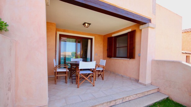 A covered patio with a table and chairs, a glass door leading to a room, and a small outdoor area with steps. - Casa Susy (Loiri Porto San Paolo)