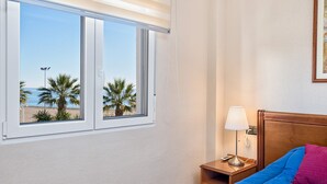 Hotelkamer met een bed, nachtkastje, lamp en raam met uitzicht op palmbomen en het strand.