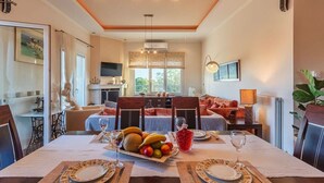 Un coin repas avec une longue table dressée pour deux personnes, une assiette de fruits et une boisson rouge.