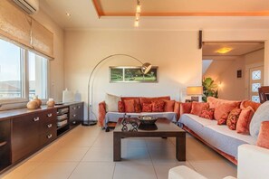 Een moderne woonkamer met een bank, salontafel en een kast. Aan de muur hangt een schilderij en er is een raam met zonwering.