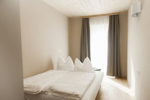 Une chambre à coucher avec un lit, du linge de lit blanc, une fenêtre avec des rideaux et un luminaire fixé au mur.