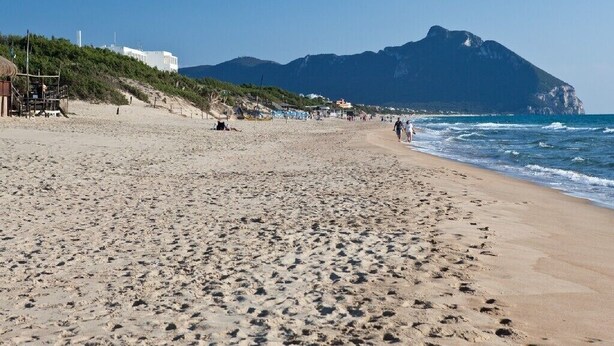 A wide sandy beach with footprints, a mountain in the background, and a few buildings on the left.
