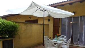 An outdoor patio with a table and chairs under a white umbrella, a potted plant, and a yellow wall.