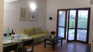 A dining area with a table set for two, a sofa, and a small table with a vase of flowers. There are framed pictures on the wall and a large sliding glass door leading to an outdoor area.