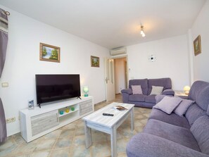 Una sala de estar con un televisor de pantalla plana, una mesa de centro blanca, un sofá púrpura y una mesita de noche con una lámpara.